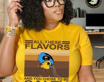 May include: A yellow crew neck t-shirt with the slogan "ALL THESE FLAVORS AND YOU CHOSE TO BE SALTY." The graphic shows a cartoon figure under a blue umbrella, sprinkling salt. The shirt is a vibrant yellow.