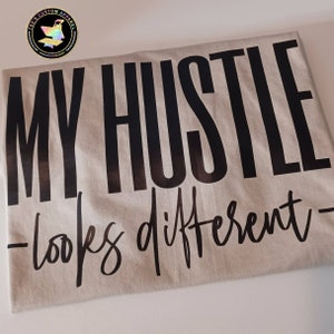 May include: A beige t-shirt with black text that reads "MY HUSTLE - looks different - "