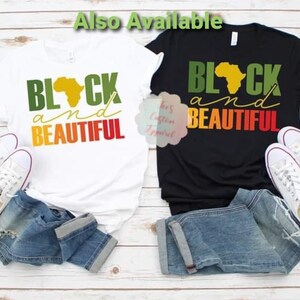 May include: Two t-shirts with the text "Black and Beautiful" in green, yellow, orange, and red. The text is overlaid on a silhouette of the continent of Africa. One t-shirt is white and the other is black.