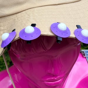 May include: A beige sun hat adorned with purple UFO-shaped clips with white domes. The hat is on a pink mannequin wearing denim overalls. A beige string with a black ring is around the mannequin's neck.