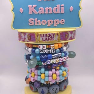 Könnte beinhalten: Ein Ausstellungsstand für 'The Kandi Shoppe' mit einem blauen Schild und rosa Text. Der Ständer ist mit bunten Perlenarmbändern verziert, von denen einige Wörter buchstabieren. Die Basis ist gelb, und die Armbänder sind gestapelt, um eine lebendige, auffällige Anzeige zu erstellen.