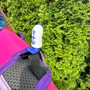Puede incluir: Un accesorio blanco en forma de botella con un dise&ntilde;o de ancla azul, sujeto a una mochila. El accesorio se asegura con un clip azul y una correa negra. La mochila es rosa con ribete morado y un panel de malla negro. Follaje verde en el fondo.