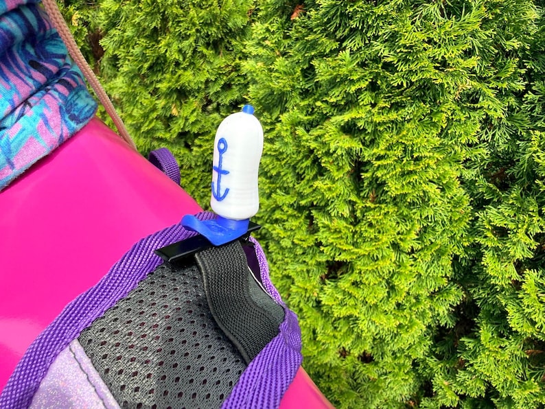 Puede incluir: Un accesorio blanco en forma de botella con un dise&ntilde;o de ancla azul, sujeto a una mochila. El accesorio se asegura con un clip azul y una correa negra. La mochila es rosa con ribete morado y un panel de malla negro. Follaje verde en el fondo.