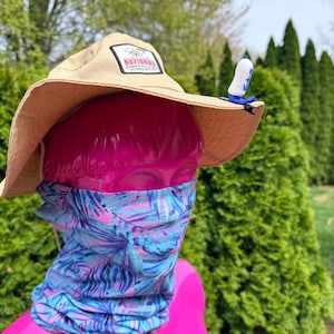 Puede incluir: Un sombrero para el sol de color beige con un parche "NATIONAL CONEY ISLAND", un accesorio azul y blanco con clip y un polaina para el cuello con estampado tropical azul y rosa. El sombrero est&aacute; sobre una cabeza de maniqu&iacute; rosa.