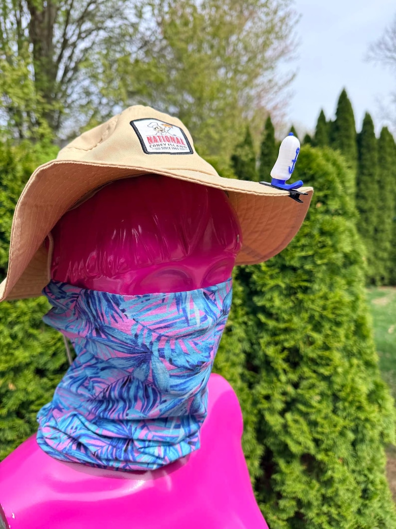 Puede incluir: Un sombrero para el sol de color beige con un parche "NATIONAL CONEY ISLAND", un accesorio azul y blanco con clip y un polaina para el cuello con estampado tropical azul y rosa. El sombrero est&aacute; sobre una cabeza de maniqu&iacute; rosa.