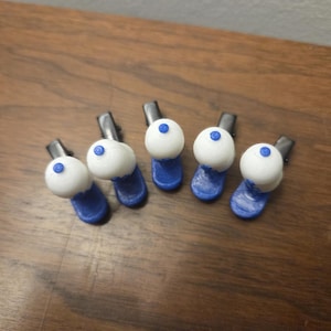 Puede incluir: Cinco pinzas para el pelo hechas a mano. Cada pinza presenta un dise&ntilde;o esf&eacute;rico blanco con un peque&ntilde;o punto azul en la parte superior, sobre una base azul. Las pinzas est&aacute;n dispuestas sobre una superficie de madera, mostrando su estilo art&iacute;stico &uacute;nico.