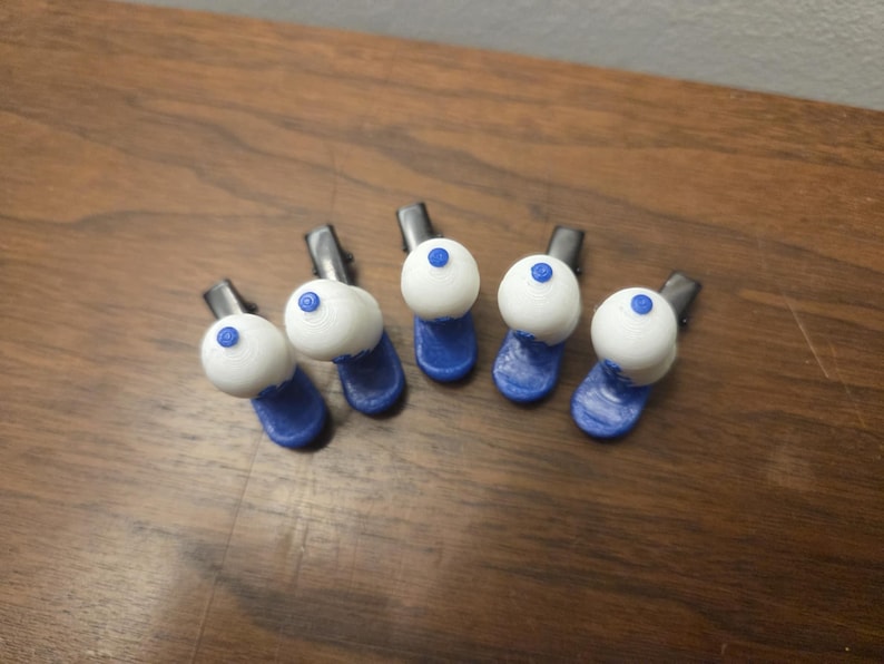 Puede incluir: Cinco pinzas para el pelo hechas a mano. Cada pinza presenta un dise&ntilde;o esf&eacute;rico blanco con un peque&ntilde;o punto azul en la parte superior, sobre una base azul. Las pinzas est&aacute;n dispuestas sobre una superficie de madera, mostrando su estilo art&iacute;stico &uacute;nico.
