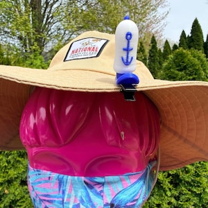 Puede incluir: Un sombrero para el sol de color beige con ala ancha, con un parche que dice "National Coney Island". Un silbato azul y blanco con dise&ntilde;o de ancla est&aacute; sujeto al sombrero. El sombrero est&aacute; sobre una forma rosa, con una mascarilla estampada.