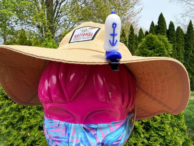 Puede incluir: Un sombrero para el sol de color beige con ala ancha, con un parche que dice "National Coney Island". Un silbato azul y blanco con dise&ntilde;o de ancla est&aacute; sujeto al sombrero. El sombrero est&aacute; sobre una forma rosa, con una mascarilla estampada.