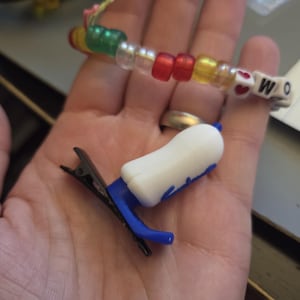Puede incluir: Una pulsera de cuentas con cuentas de colores que deletrean "WO" y una peque&ntilde;a pinza para el pelo blanca y azul. La pulsera presenta cuentas rojas, amarillas, verdes y transparentes. La pinza para el pelo tiene una parte superior blanca y detalles azules.