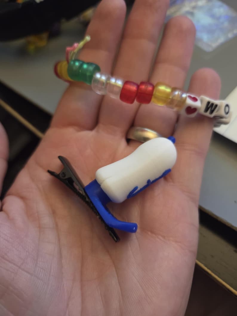 Puede incluir: Una pulsera de cuentas con cuentas de colores que deletrean "WO" y una peque&ntilde;a pinza para el pelo blanca y azul. La pulsera presenta cuentas rojas, amarillas, verdes y transparentes. La pinza para el pelo tiene una parte superior blanca y detalles azules.