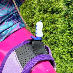 Puede incluir: Un tap&oacute;n de botella blanco y azul con un dise&ntilde;o de ancla azul. El tap&oacute;n est&aacute; sujeto a un arn&eacute;s rosa y morado con una secci&oacute;n de malla negra. En el fondo se ve una tela estampada azul, morada y rosa.