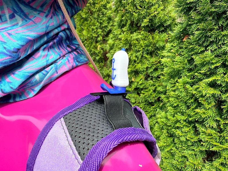 Puede incluir: Un tap&oacute;n de botella blanco y azul con un dise&ntilde;o de ancla azul. El tap&oacute;n est&aacute; sujeto a un arn&eacute;s rosa y morado con una secci&oacute;n de malla negra. En el fondo se ve una tela estampada azul, morada y rosa.