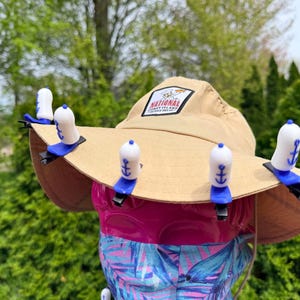 Puede incluir: Un sombrero de sol beige con ala ancha, con un parche que dice "National Coney Island". El sombrero est&aacute; adornado con varios clips para sombrero blancos y azules en forma de ancla. El sombrero se muestra sobre una superficie rosa y estampada.