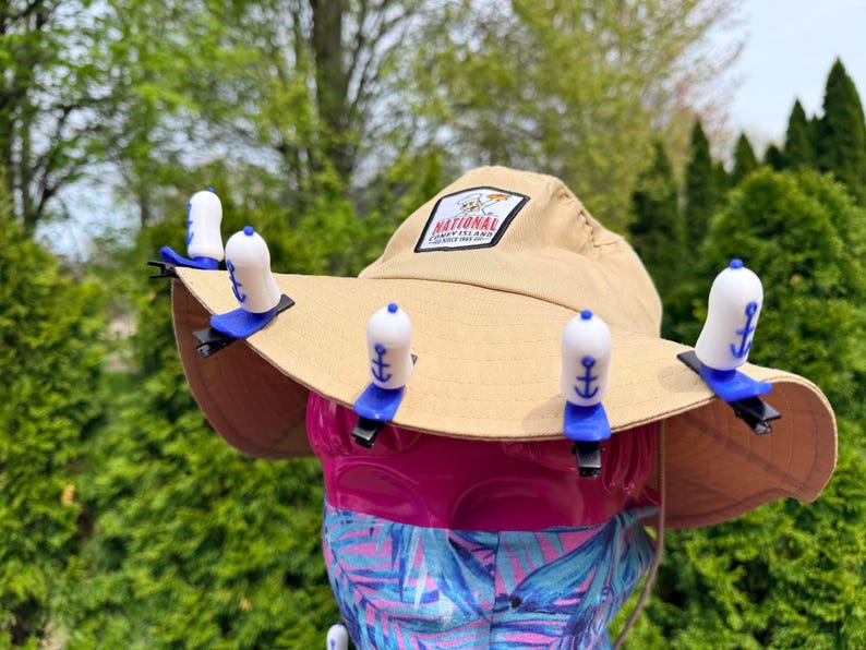 Puede incluir: Un sombrero de sol beige con ala ancha, con un parche que dice "National Coney Island". El sombrero est&aacute; adornado con varios clips para sombrero blancos y azules en forma de ancla. El sombrero se muestra sobre una superficie rosa y estampada.
