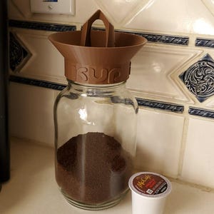 May include: A glass jar filled with ground coffee, topped with a brown funnel-shaped lid with a handle. A single white and brown McCafe K-cup sits next to the jar. The jar is on a white countertop.