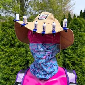 Puede incluir: Un sombrero para el sol de color beige con un parche de "National Park", adornado con clips blancos y azules en forma de ancla. Una cabeza de maniqu&iacute; rosa lleva un cuello con estampado azul y morado y una mochila morada.