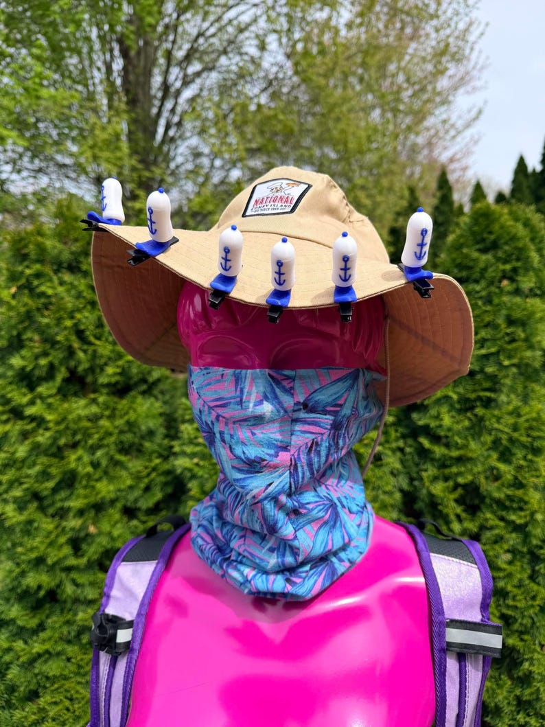 Puede incluir: Un sombrero para el sol de color beige con un parche de "National Park", adornado con clips blancos y azules en forma de ancla. Una cabeza de maniqu&iacute; rosa lleva un cuello con estampado azul y morado y una mochila morada.
