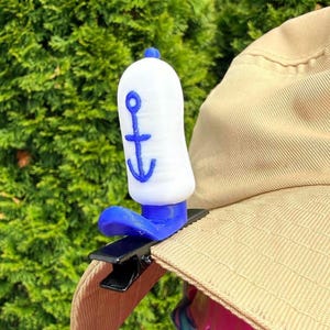 Könnte beinhalten: Ein weiß-blauer Hutclip im nautischen Stil mit Ankerdesign. Der Clip ist an einem beigefarbenen Sonnenhut befestigt. Der Clip besteht aus Kunststoff und hat eine schwarze Metallklammer.