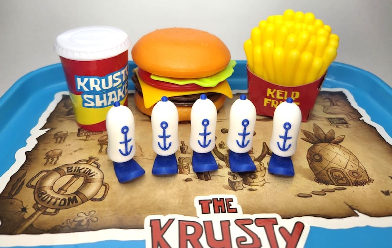 Puede incluir: Un juego de comida de juguete colorido con una hamburguesa, papas fritas, una bebida y cinco sombreros de marinero blancos y azules con dise&ntilde;os de anclas. El juego est&aacute; en una bandeja con un dise&ntilde;o de mapa y el texto "The Krusty".