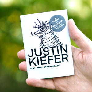 Könnte beinhalten: Eine weiße Karte mit einer schwarzen Illustration eines wurmartigen Wesens und dem Namen "JUSTIN KIEFER" in fetten schwarzen Buchstaben. Der Text "War mein Mitbewohner!" steht unter dem Namen. Ein blauer Kreis mit deutschem Text befindet sich oben rechts.
