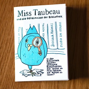 Könnte beinhalten: Ein kleines, illustriertes Buch mit dem Titel "Miss Taubeau und die Geheimnisse der Bibliothek", das einen blauen Vogel mit einer Lupe zeigt. Das Cover enthält deutschen Text und eine Cartoon-Illustration eines Vogeldetektivs. Das Buch liegt auf einer Holzoberfläche.