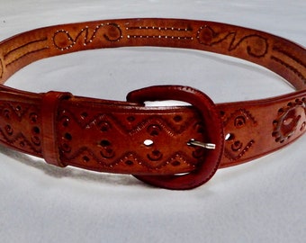 May include: A brown leather belt with an intricate carved design and a silver buckle. The belt is decorated with a repeating pattern of swirls and lines.