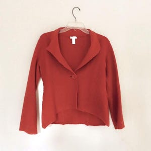 May include: A rust-colored blazer with a single-button closure and a wide collar. The jacket is hanging on a clear plastic hanger against a white background. The blazer has long sleeves and a slightly asymmetrical hemline.