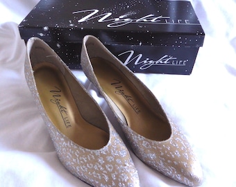 May include: A pair of pointed-toe pumps with a beige base and a silver glitter leopard print pattern. The interior soles are gold with the text Night Life printed in black. The shoes are positioned in front of a black box labeled Night Life.