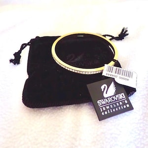 Puede incluir: Un brazalete dorado con una fila de pequeños cristales transparentes. El brazalete descansa sobre una bolsa de terciopelo negro, con una etiqueta que dice "Swarovski Jeweler's collection". Es una joya.