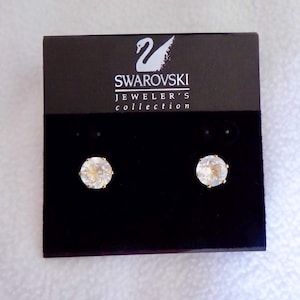 Peut inclure: Une paire de boucles d'oreilles Swarovski sur une carte d'exposition noire. Les boucles d'oreilles présentent des cristaux ronds et transparents sertis dans du métal doré. Le logo Swarovski est visible sur la carte.