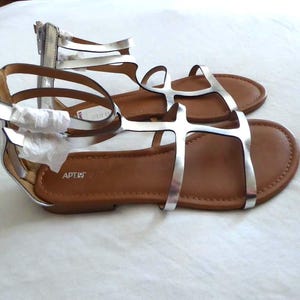 Può includere: Un paio di sandali bassi con cinturini color argento metallizzato in stile gladiatore e chiusura con zip sul retro. Il plantare è color cuoio con il nome del marchio Apt. 9 stampato in bianco. I cinturini presentano carta protettiva bianca.