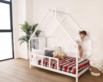 以下が含まれることがあります： 赤と黒のチェック柄のマットレスが付いた白い木製の家の形のベッドフレーム。若い女の子がベッドフレームの上に立って、下を見ている。