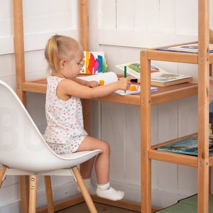 May include: A young girl sits at a wooden desk with a white chair, coloring in a notebook. The desk is part of a wooden shelving unit with books on the shelves.