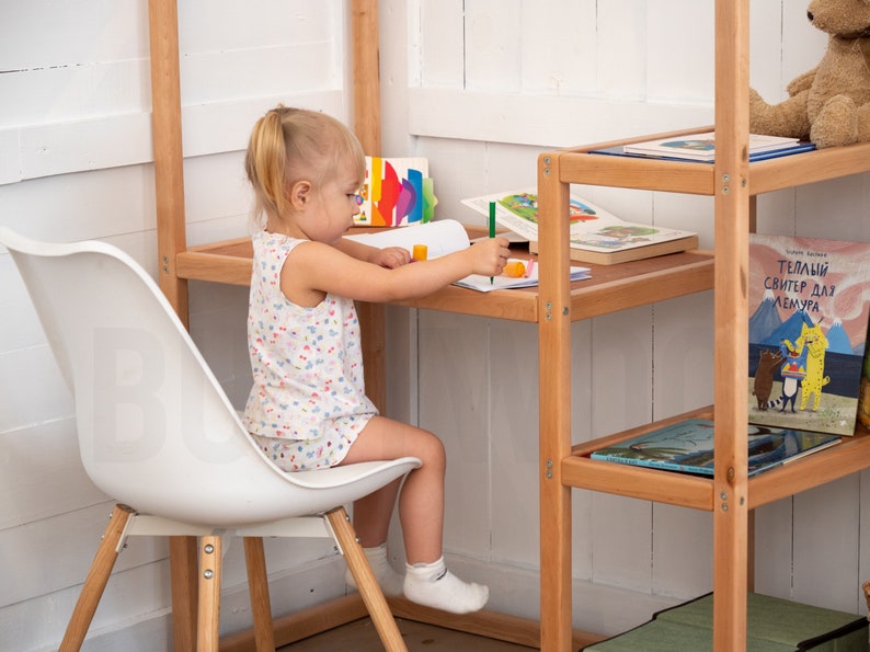 May include: A young girl sits at a wooden desk with a white chair, coloring in a notebook. The desk is part of a wooden shelving unit with books on the shelves.