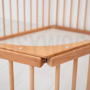 Pu&ograve; includere: Primo piano di un angolo di un box per bambini in legno con una ringhiera superiore curva e viti in metallo. Il box &egrave; realizzato in legno naturale e ha una finitura marrone chiaro.