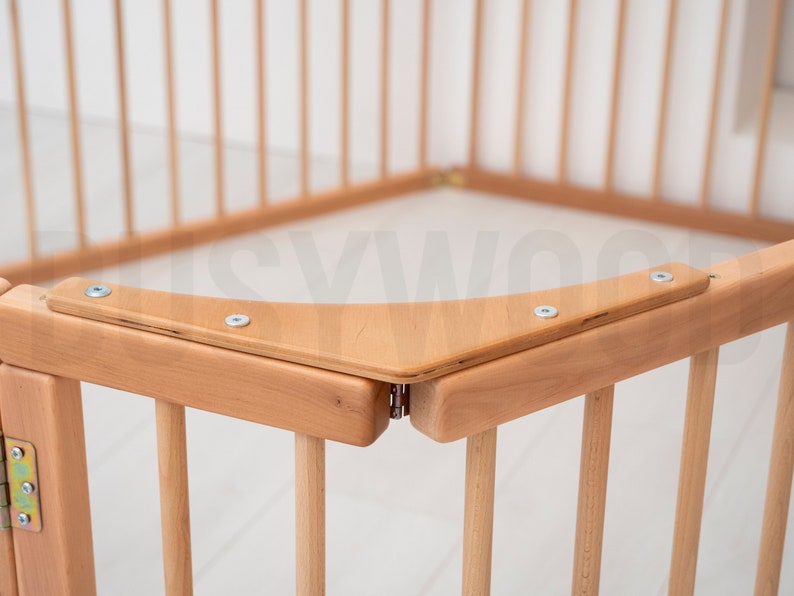 Pu&ograve; includere: Primo piano di un angolo di un box per bambini in legno con una ringhiera superiore curva e viti in metallo. Il box &egrave; realizzato in legno naturale e ha una finitura marrone chiaro.