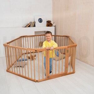 Pu&ograve; includere: Un recinto di gioco esagonale in legno con un bambino all'interno. Il recinto &egrave; fatto di legno naturale. Il bambino indossa una camicia gialla e guarda la fotocamera.