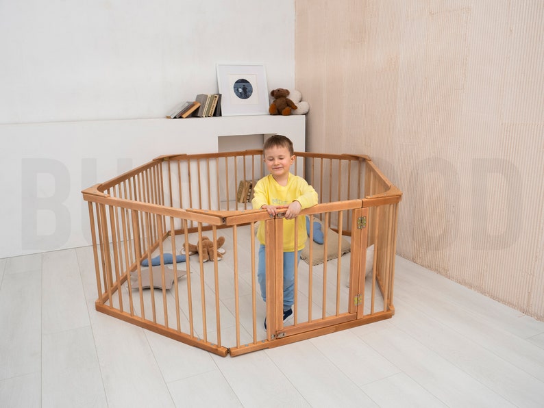 Pu&ograve; includere: Un recinto di gioco esagonale in legno con un bambino all'interno. Il recinto &egrave; fatto di legno naturale. Il bambino indossa una camicia gialla e guarda la fotocamera.