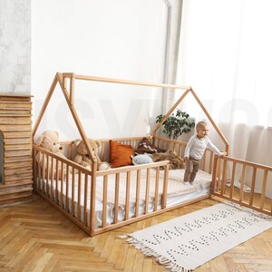 May include: A natural wood-framed house bed with a slatted side and a gabled roof. The bed is furnished with a mattress, pillows, and stuffed animals. A small child stands near the open gate. A patterned rug is on the floor.