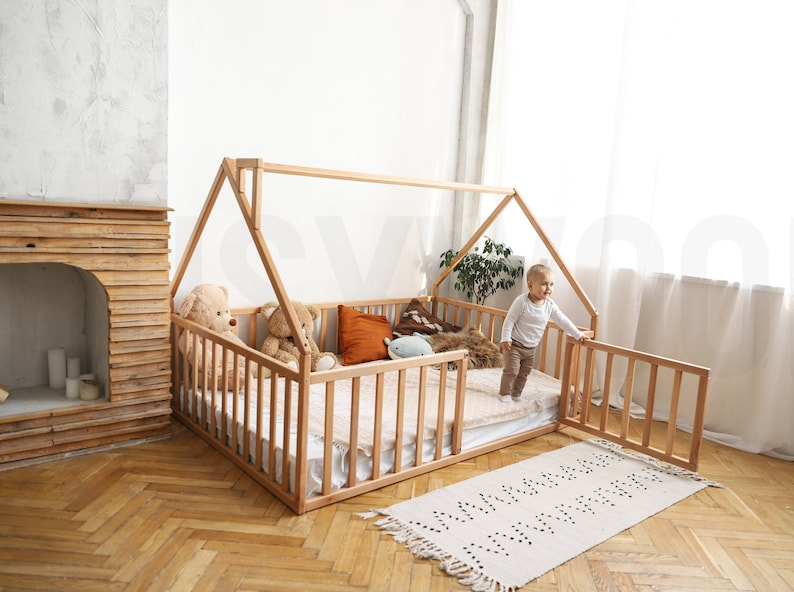 May include: A natural wood-framed house bed with a slatted side and a gabled roof. The bed is furnished with a mattress, pillows, and stuffed animals. A small child stands near the open gate. A patterned rug is on the floor.