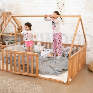 May include: A wooden house-shaped bed frame with a built-in bookshelf. The bed has a low profile with a slatted side rail. Two children are playing on the bed. The bed is filled with pillows, a blanket, and a white sheet.