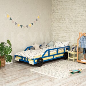 K&ouml;nnte beinhalten: Ein blaues und gelbes Spielzeugauto-Bettgestell mit einer wei&szlig;en und gelben Bettw&auml;sche. Das Bett steht in einem Zimmer mit einer wei&szlig;en Backsteinwand, einer gr&uuml;nen Pflanze und einem Holz-Kleiderst&auml;nder.