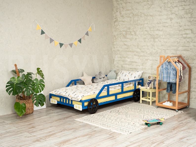 K&ouml;nnte beinhalten: Ein blaues und gelbes Spielzeugauto-Bettgestell mit einer wei&szlig;en und gelben Bettw&auml;sche. Das Bett steht in einem Zimmer mit einer wei&szlig;en Backsteinwand, einer gr&uuml;nen Pflanze und einem Holz-Kleiderst&auml;nder.