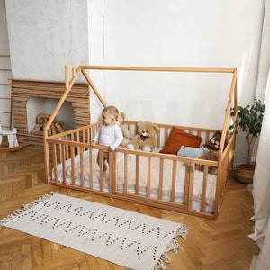 May include: A light-colored wooden house-shaped bed frame with a slatted design. The bed is furnished with a mattress, pillows, and stuffed animals. A small child stands inside the bed. A rug with a zigzag pattern is on the floor.