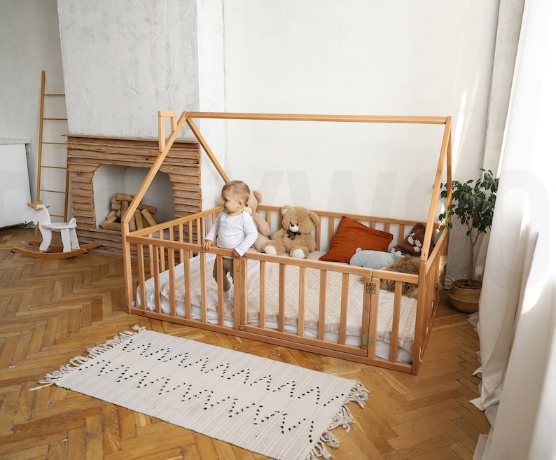 May include: A light-colored wooden house-shaped bed frame with a slatted design. The bed is furnished with a mattress, pillows, and stuffed animals. A small child stands inside the bed. A rug with a zigzag pattern is on the floor.
