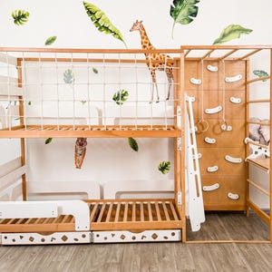 Pu&ograve; includere: Un letto a castello in legno con una parete da arrampicata, uno scivolo e sbarre per scimmie. Il letto ha una finitura in legno naturale e un cassetto bianco. Il muro &egrave; decorato con decalcomanie di animali e foglie.