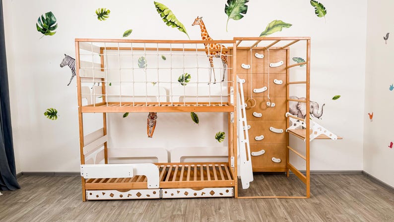 Pu&ograve; includere: Un letto a castello in legno con una parete da arrampicata, uno scivolo e sbarre per scimmie. Il letto ha una finitura in legno naturale e un cassetto bianco. Il muro &egrave; decorato con decalcomanie di animali e foglie.