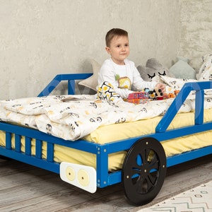 K&ouml;nnte beinhalten: Ein blaues Holzbett in Form eines Autos mit einer gelben Matratze und einem Kind, das darauf sitzt und mit Spielzeug spielt. Das Bett hat schwarze R&auml;der und wei&szlig;e Scheinwerfer.