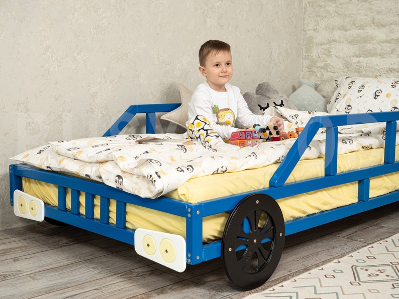 K&ouml;nnte beinhalten: Ein blaues Holzbett in Form eines Autos mit einer gelben Matratze und einem Kind, das darauf sitzt und mit Spielzeug spielt. Das Bett hat schwarze R&auml;der und wei&szlig;e Scheinwerfer.